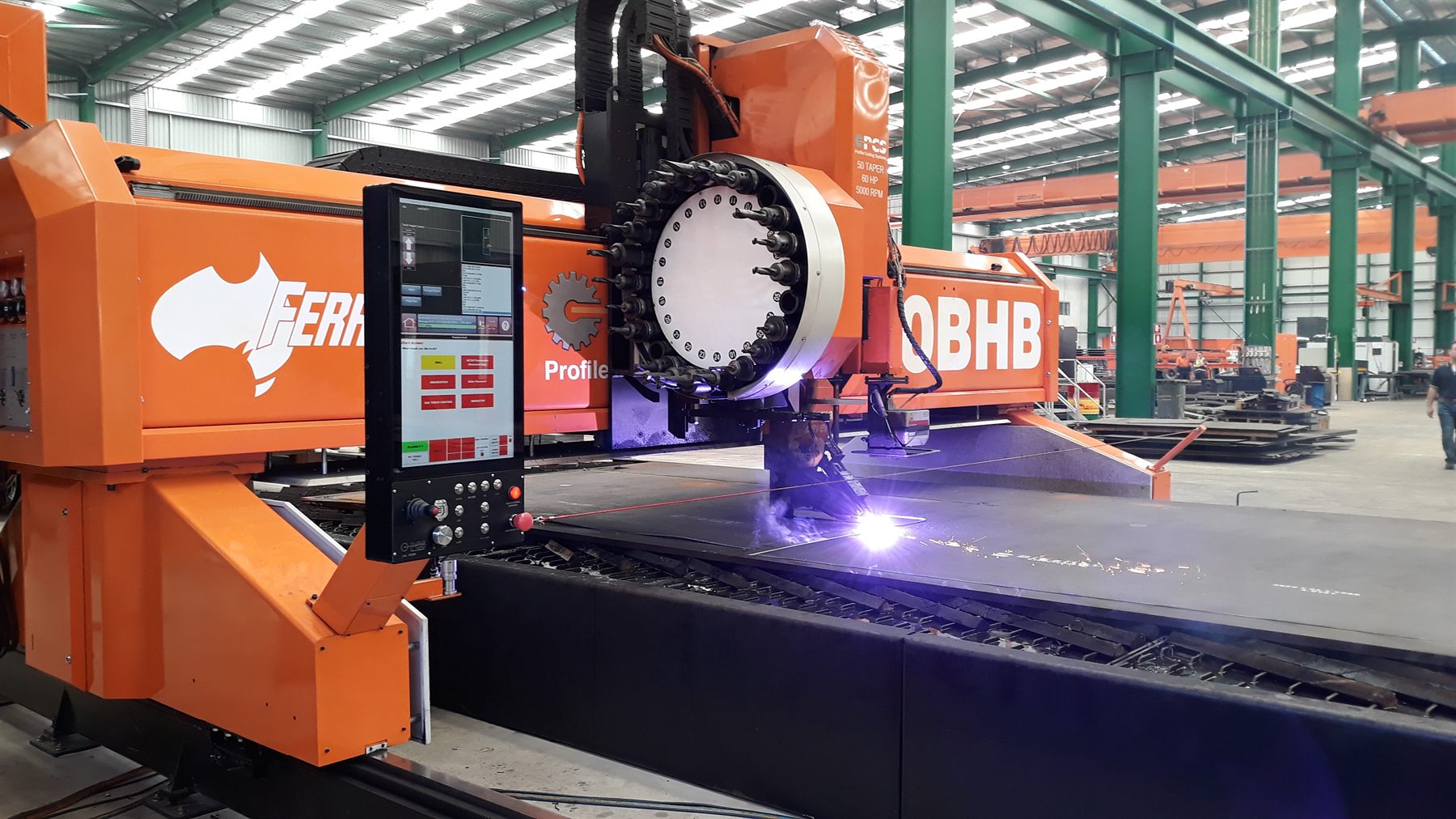 A large orange CNC gantry profile cutting machine with a multi-tool drill head striking a plasma arc on heavy steel plate in a fabrication workshop.